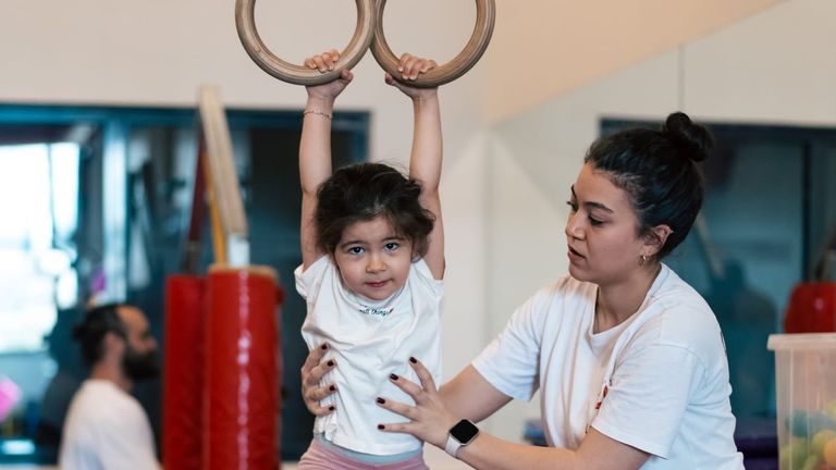 Eğitmen çocuğu belinden tutarken çocuk jimnastik halkalarına asılıyor